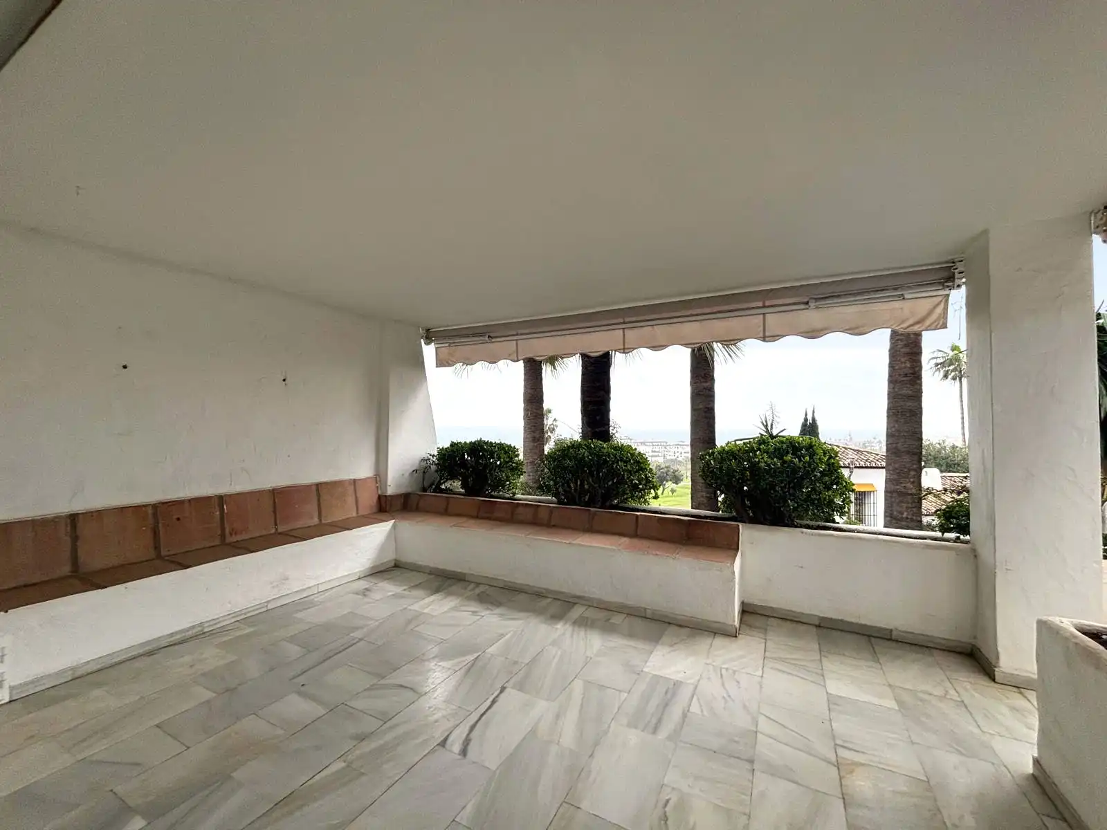 Covered terrace with built-in benches and open views over the garden, pool and surroundings of Brisas II, Manilva; the sea can be seen in the distance in this image.